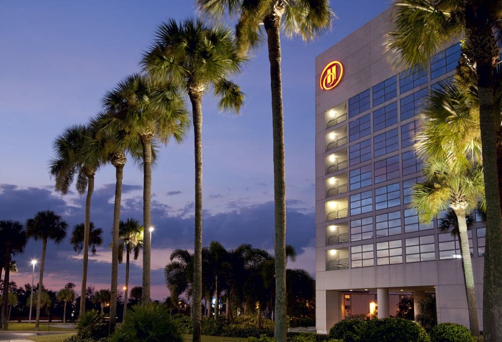 Exterior of Hilton Rialto Place in Melbourne, Florida, at dusk, with palm trees, the Hilton logo, and illuminated guestroom windows.