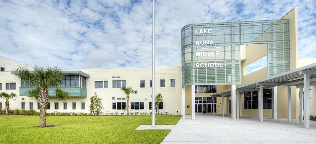 Exterior of Lake Nona Middle School in Orlando, Florida, a modern campus with a green-tinted entrance screen, covered walkway, palm trees, and a flagpole.