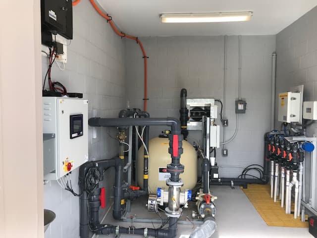 Mechanical equipment room for the West Melbourne Community Park splash pad, showing filtration piping, valves, a filter vessel, and wall-mounted controls.